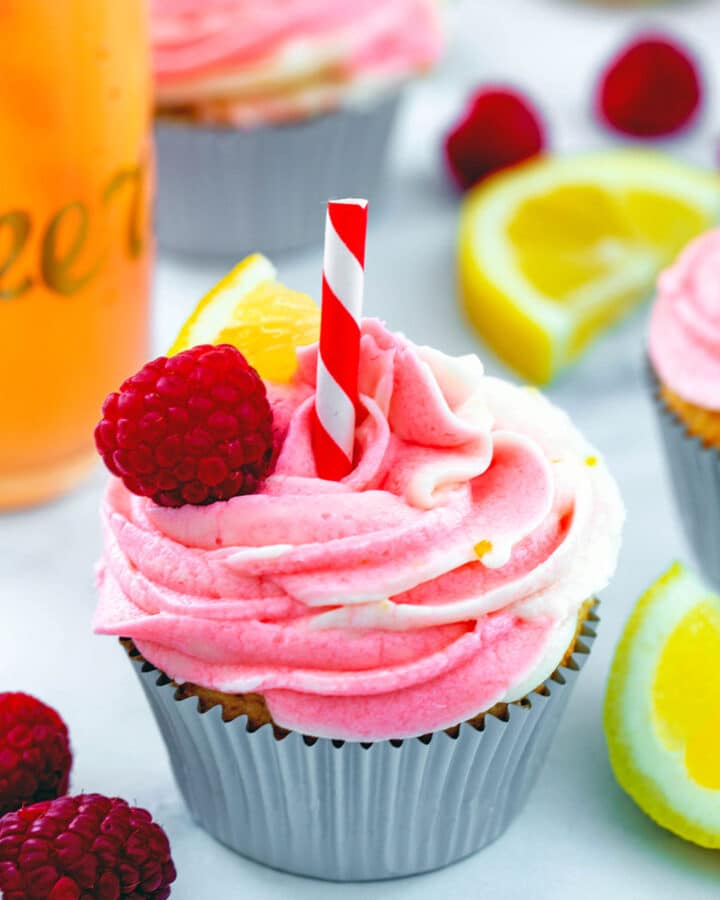 Head-on view of a raspberry lemonade cupcake with swirled frosting, straw, and raspberries and lemon wedges all around with more cupcakes in background