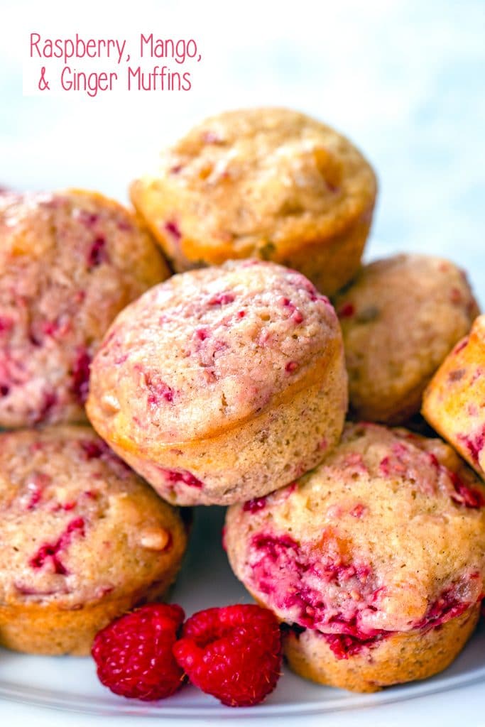 Head-on view of a plate full of raspberry, mango, ginger muffins piled up with fresh raspberries on the side and recipe title at top