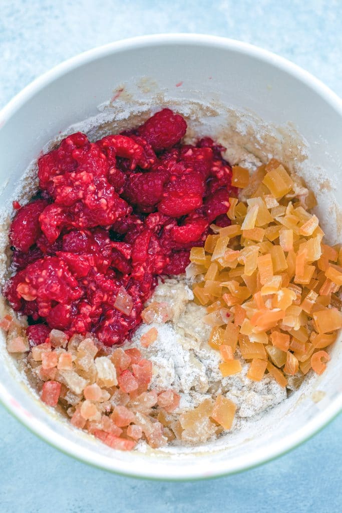 Overhead view of a mixing bowl filled with muffin batter and chopped fresh raspberries, chopped dried mango, and chopped candied ginger