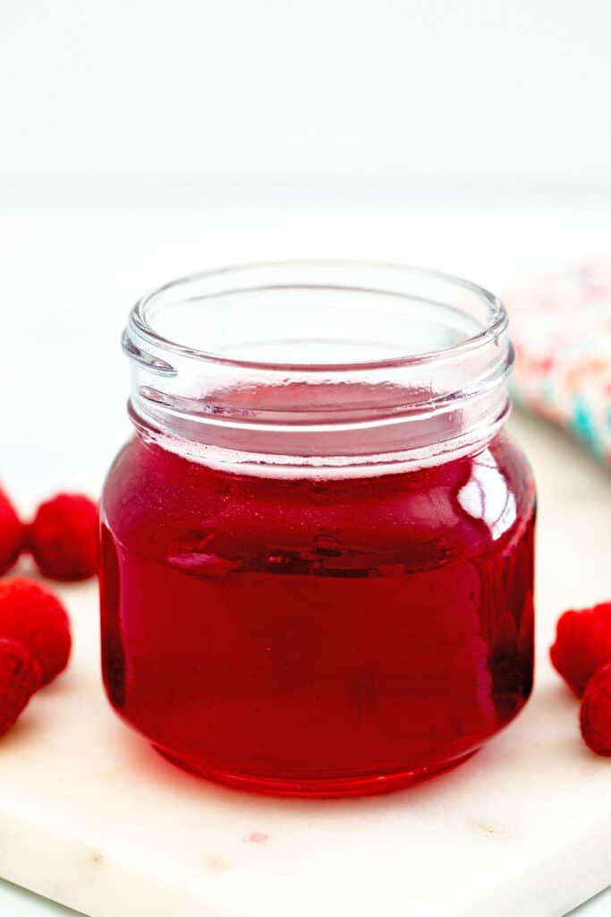 Head-on view of raspberry simple syrup in a small jar with fresh raspberries all around.
