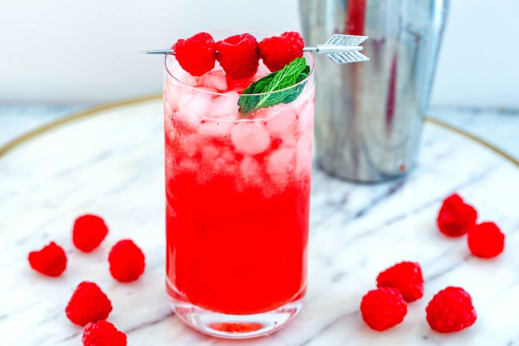 Landscape view of a raspberry whiskey cocktail with raspberry and mint garnish on a marble tray with raspberries scattered around and a cocktail shaker in the background