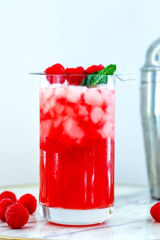 Straight-on view of a raspberry whiskey cocktail with vanilla simple syrup with raspberry mint garnish on a marble tray with raspberries and cocktail shaker in the background