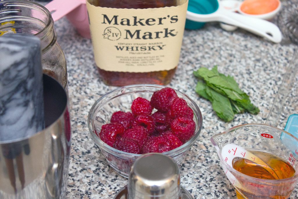 Close-up of bowl of raspberries with vanilla simple syrup, mint leaves, cocktail shaker, whisky bottle, measuring cups and spoons on the counter