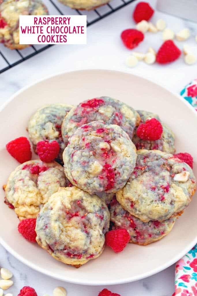 Raspberry white chocolate cookies on a plate with more cookies on rack in background and raspberries and white chocolate chips all around with recipe title at top.