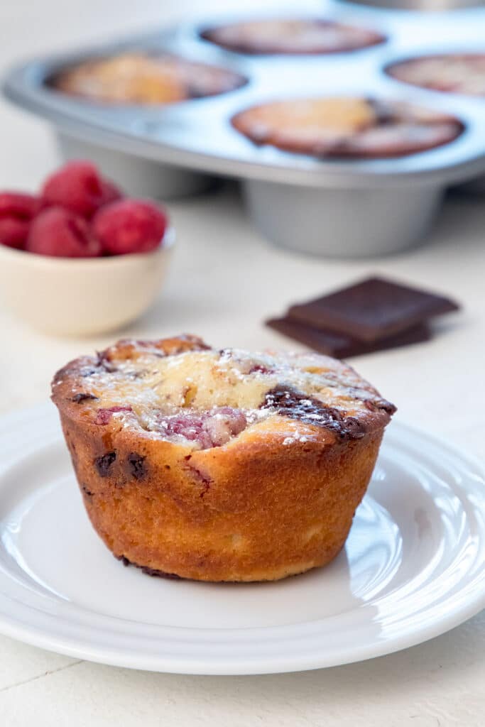 Head-on view of a raspberry dark chocolate muffin on a white plate with dark chocolate squares, bowl of raspberries, and tin with more muffins in the background.