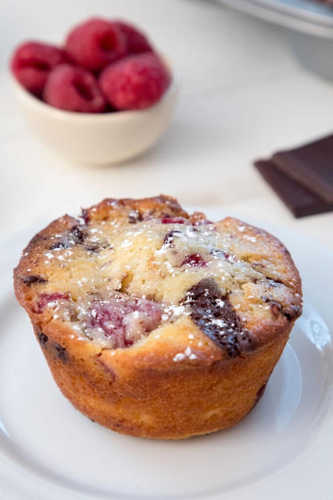 Overhead closeup view of a raspberry dark chocolate muffin with dark chocolate squares and bowl of raspberries in background.