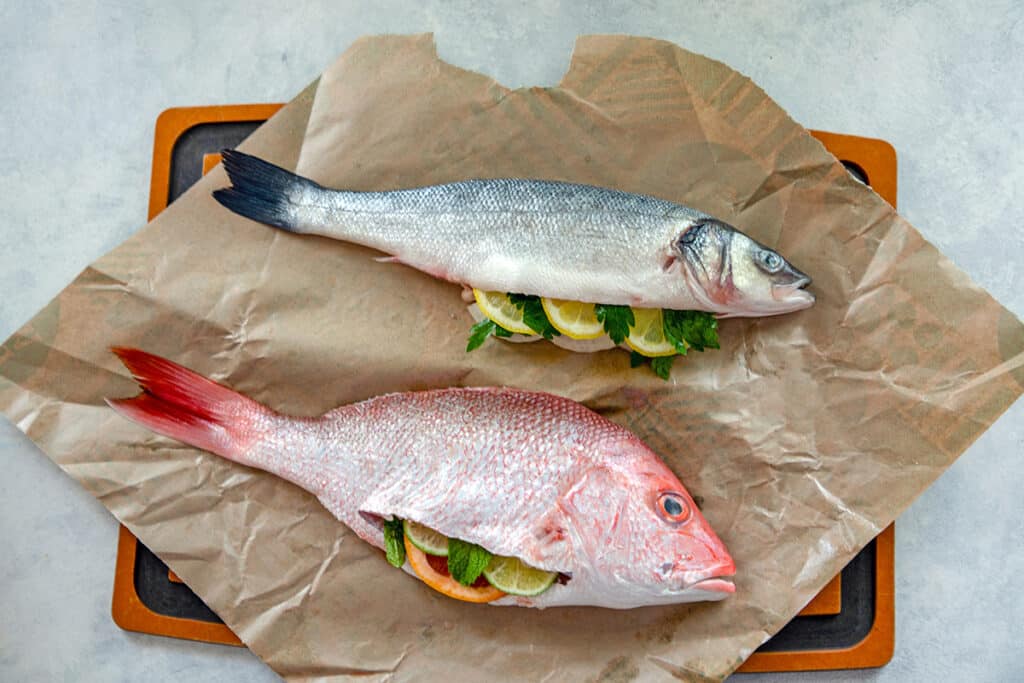Overhead view of whole branzino and whole red snapper stuffed with herbs and citrus on parchment paper.