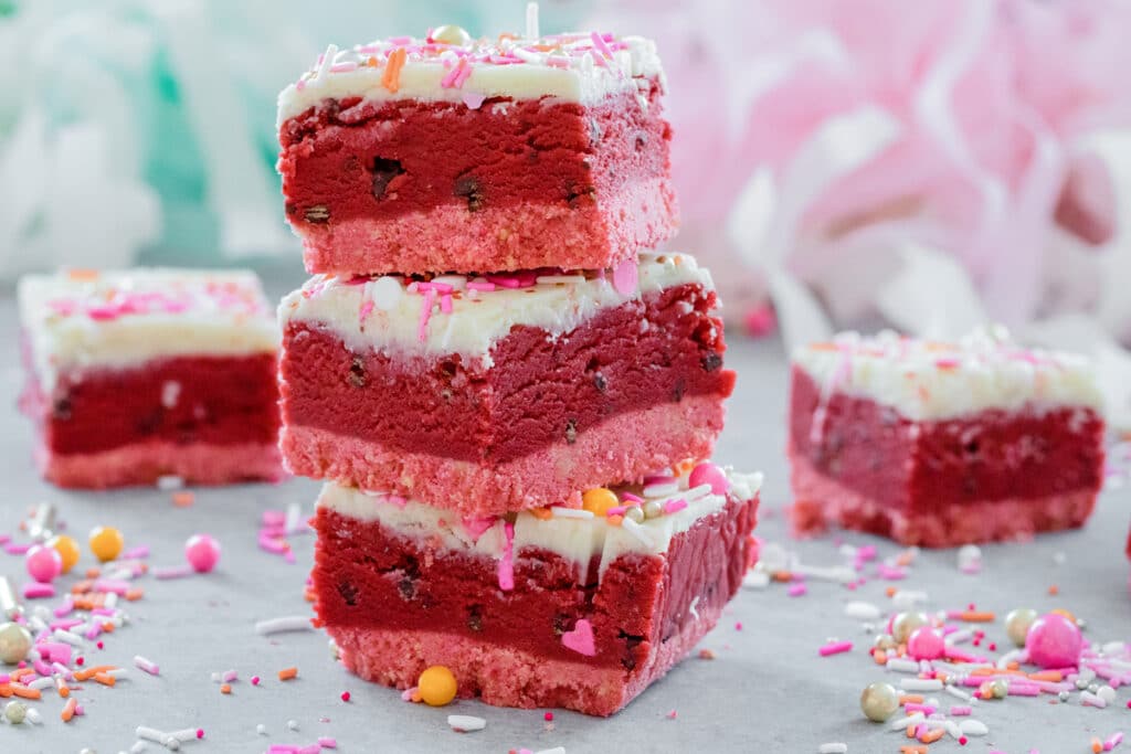 Landscape photo of stack of three red velvet cookie dough bars with white chocolate ganache and Valentine's Day sprinkles with more cookie dough bars and sprinkles all around.