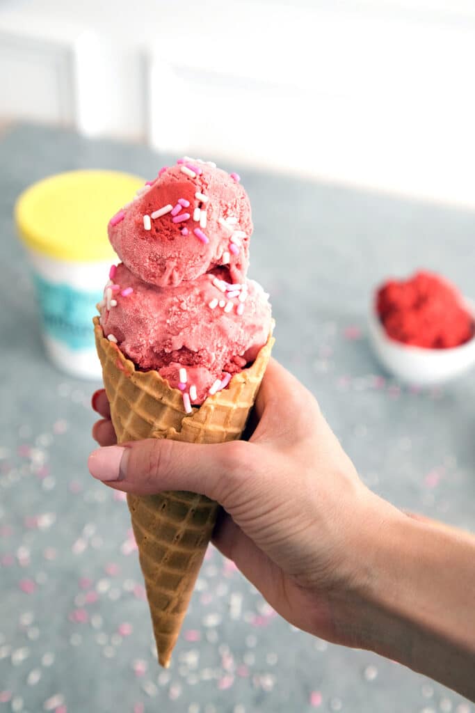 Cone of red velvet ice cream with cookie dough being held out.