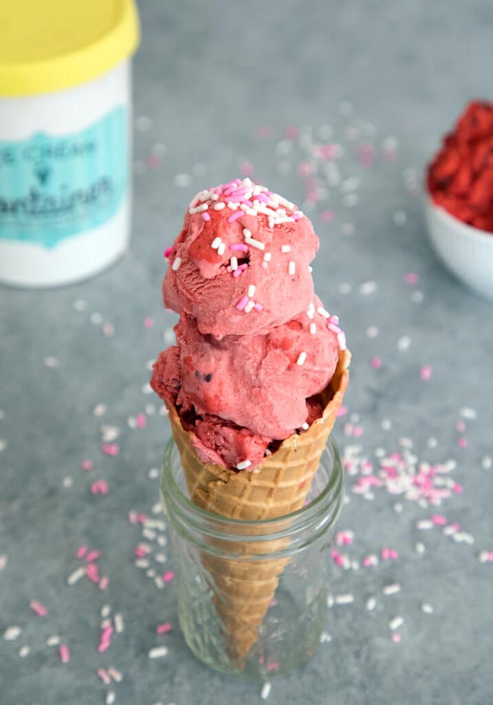 Close-up overhead photo of red velvet ice cream with cookie dough in a cone in a mason jar surrounded by sprinkles with an ice cream container and bowl of red velvet cookie dough in background.