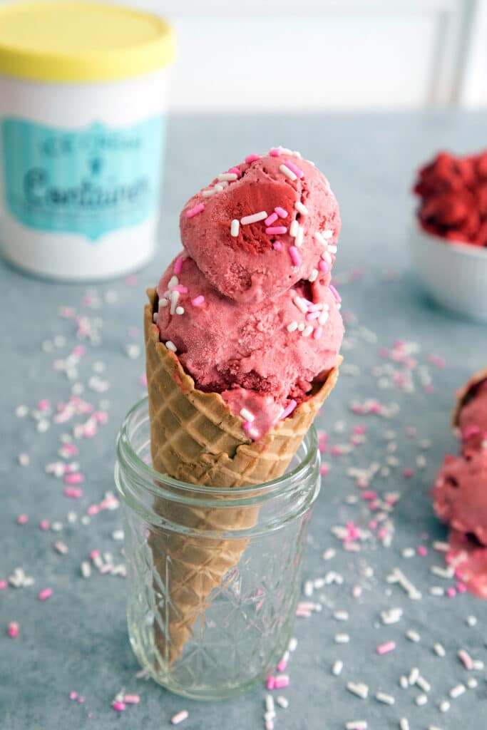 Head-on view of red velvet ice cream with cookie dough in an ice cream cone sitting in a mason jar surrounded by sprinkles and a ice cream container.