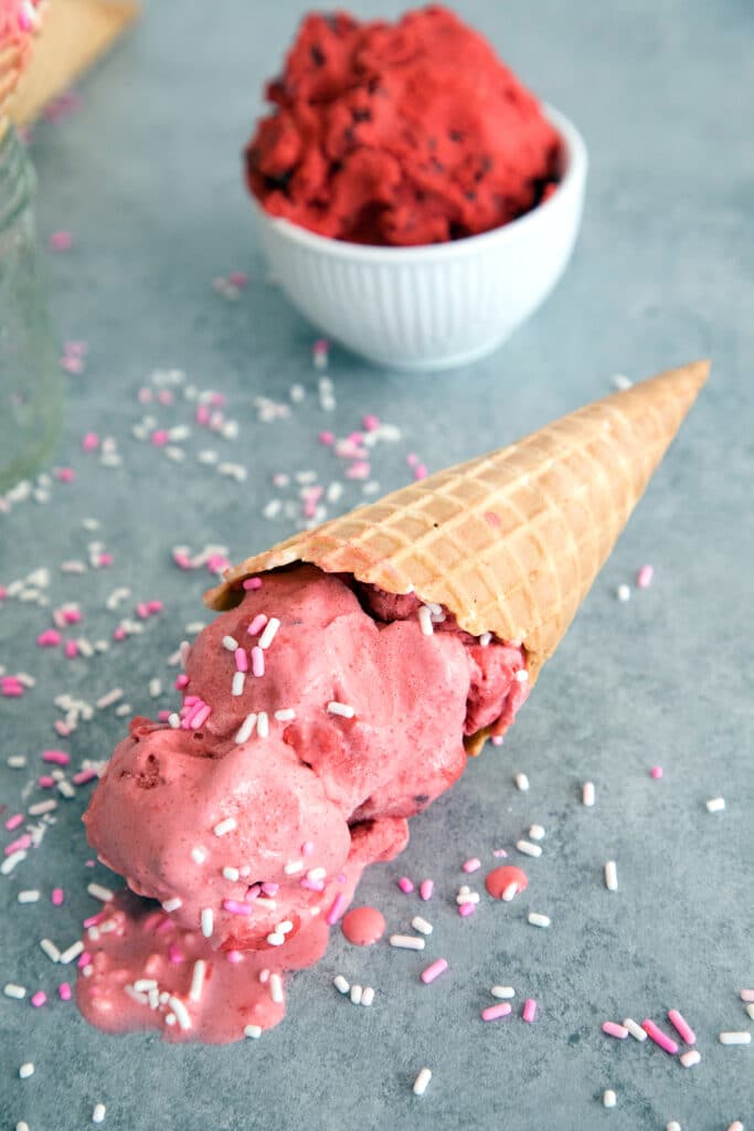 Overhead view of red velvet ice cream with cookie dough in an ice cream cone laying and melting on grey surface with bowl of red velvet cookie dough and sprinkles in background.