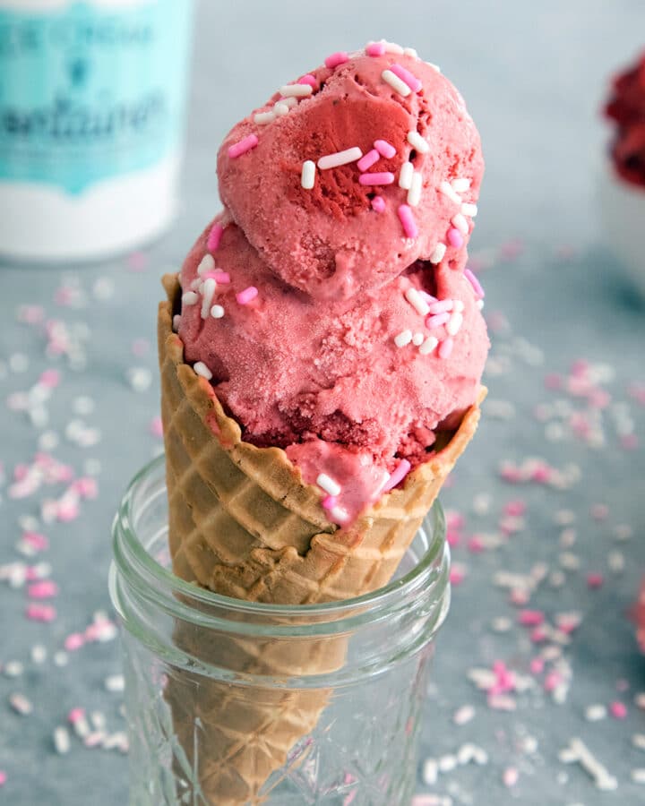 red velvet cookie dough ice cream in a cone sitting in a jar with sprinkles all around.