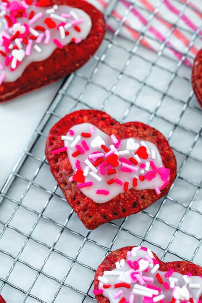 Overhead view of heart-shaped red velvet pop tart with icing and sprinkles on a baking rack with more pop tarts in background.