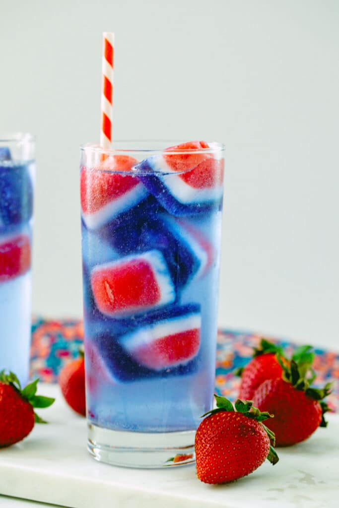 Head-on view of glass of soda water with red, white, and blue ice cubes, red and white straw, with strawberries all around