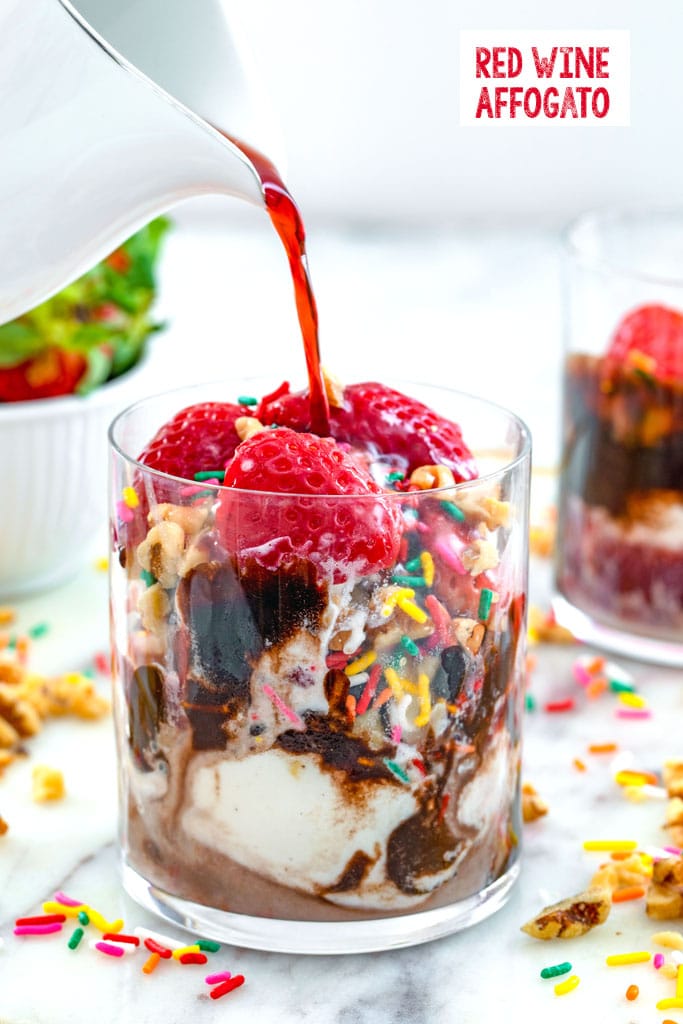 Head-on view of a glass of vanilla ice cream with chocolate, strawberries, walnuts, and sprinkles with red wine being poured over it for a red wine affogato with recipe title at top of image