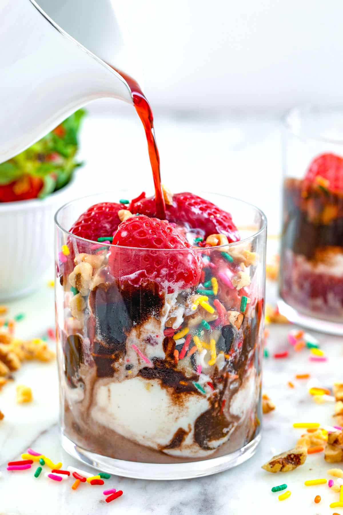Head-on view of a glass of vanilla ice cream with chocolate, strawberries, walnuts, and sprinkles with red wine being poured over it.