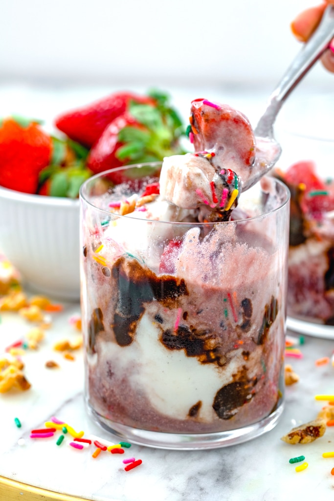 Head-on view of a red wine affogato in a glass with spoon holding ice cream, wine, strawberries, and sprinkles with more strawberries and sprinkles in background