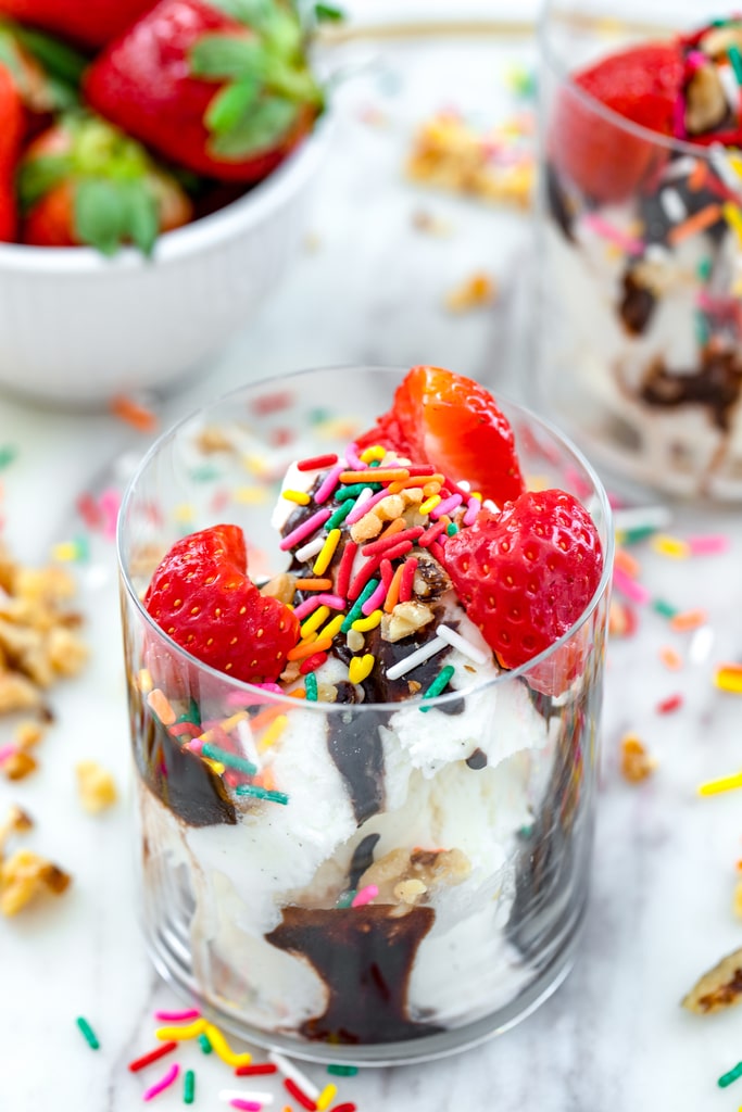 Overhead view of a glass filled with vanilla ice cream with fudge sauce, strawberries, walnuts, and sprinkles with second glass, bowl of strawberries, and sprinkles and walnuts in background