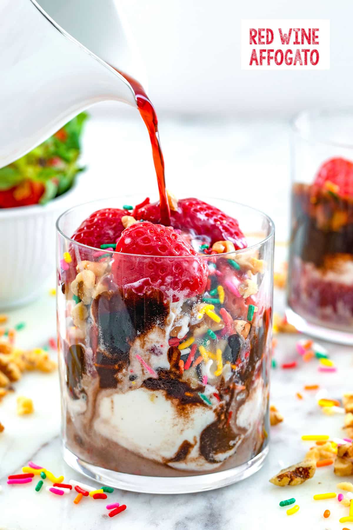Head-on view of a glass of vanilla ice cream with chocolate, strawberries, walnuts, and sprinkles with red wine being poured over it for a red wine affogato with recipe title at top of image.