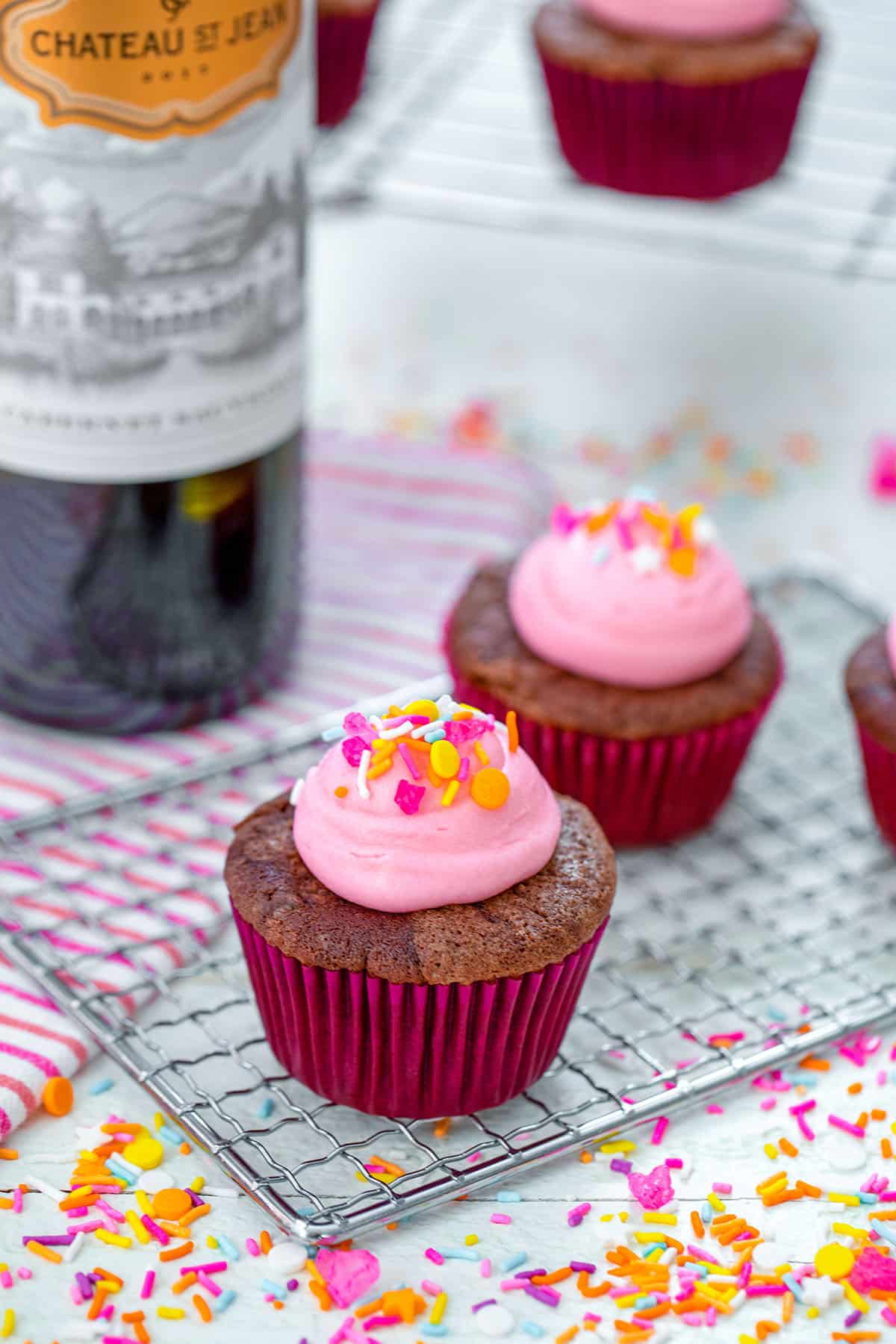 Head-on view from a distance of red wine cupcakes on a cooling rack with pink frosting and sprinkles with bottle of wine, more cupcakes, and sprinkles in background.