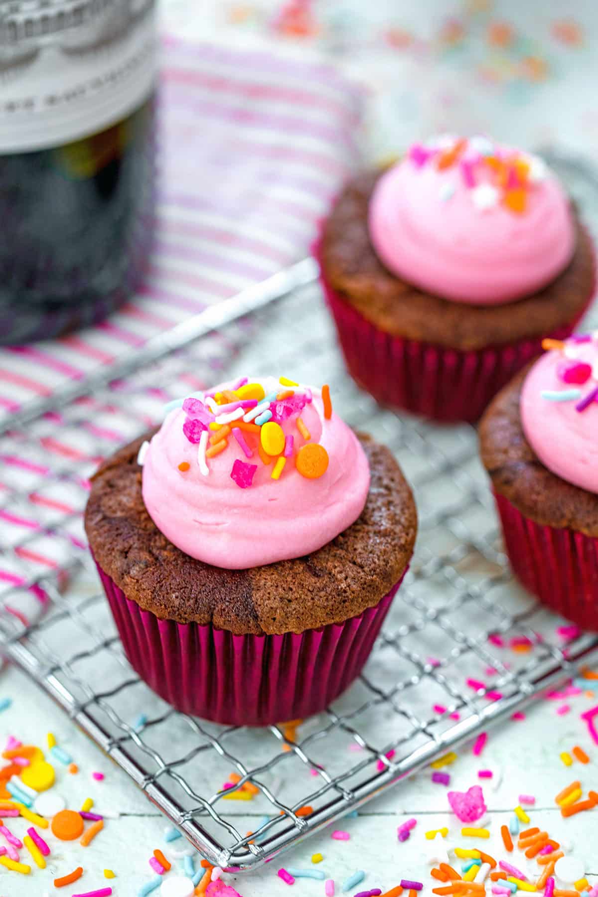 Head-on view of a red wine cupcake with pink frosting and sprinkles on a baking rack with more cupcakes and sprinkles in the background.
