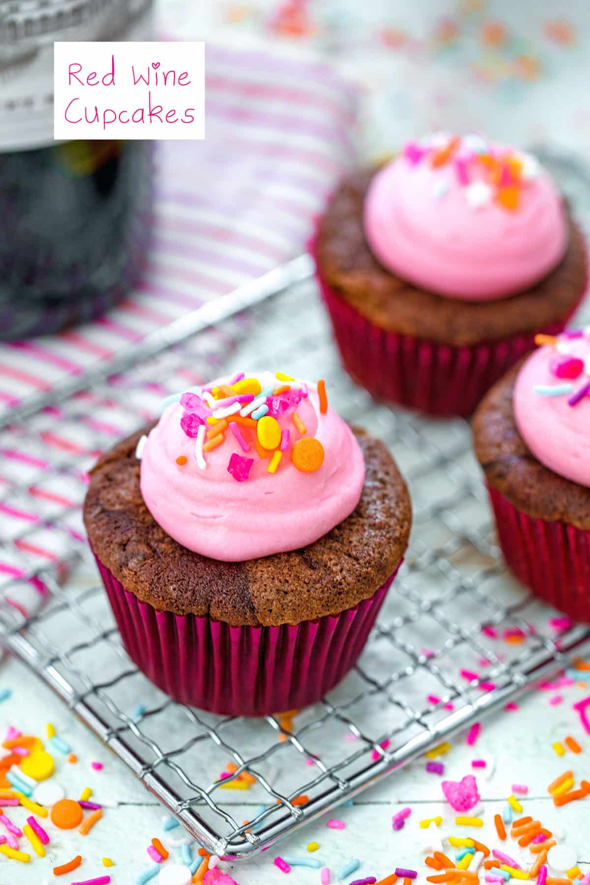 Head-on view of a red wine cupcake with pink frosting and sprinkles on a baking rack with more cupcakes and sprinkles in the background with recipe title at top.