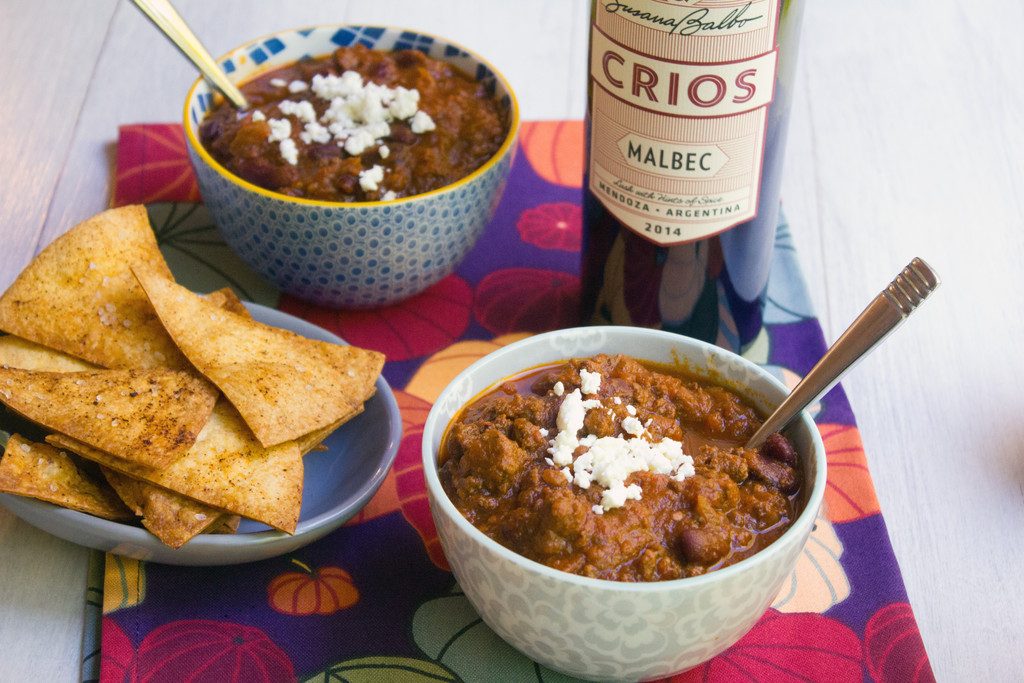 Overhead view of a bowl of red wine pumpkin chili topped with feta cheese with bottle of red wine, second bowl of chili, and homemade tortilla chips in background