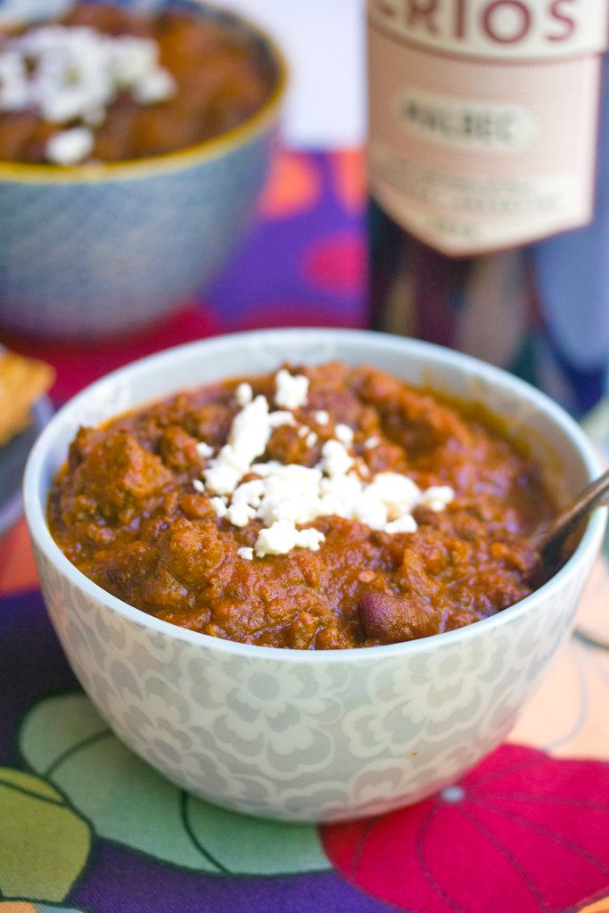 Head-on closeup view of red wine pumpkin chili topped with feta cheese with second bowl of chili and bottle of red wine in background