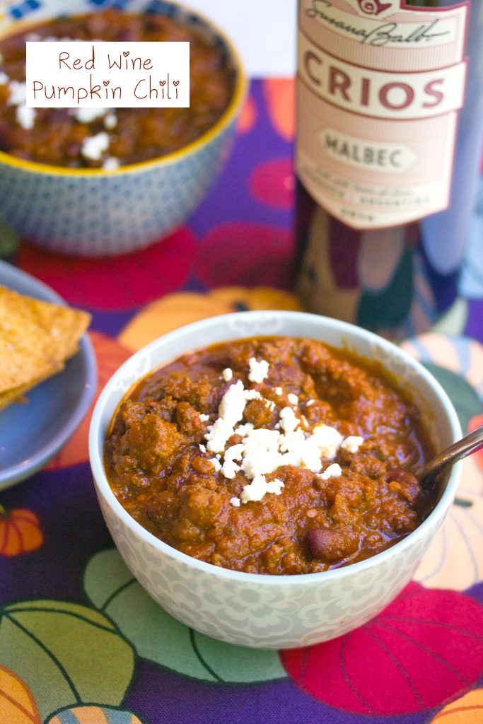 Overhead view of a bowl of red wine pumpkin chili topped with feta cheese with bottle of red wine, second bowl of chili, and homemade tortilla chips in background and recipe title at top