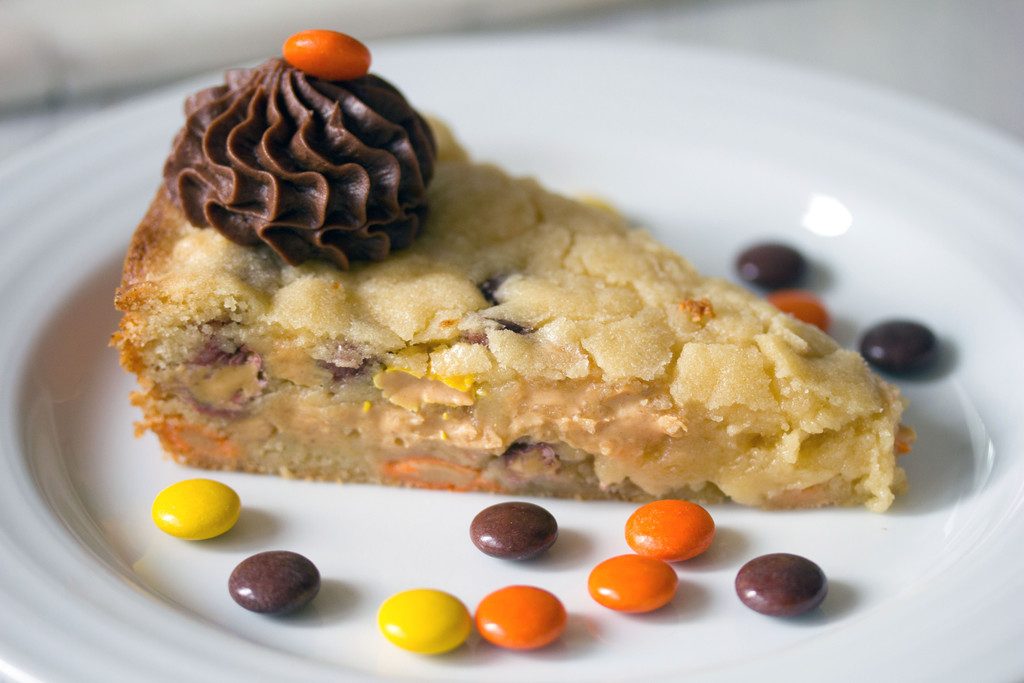 Landscape closeup head-on view of a slice of Reese's Pieces cookie cake with a dollop of chocolate frosting on a white plate surrounded by Reese's Pieces