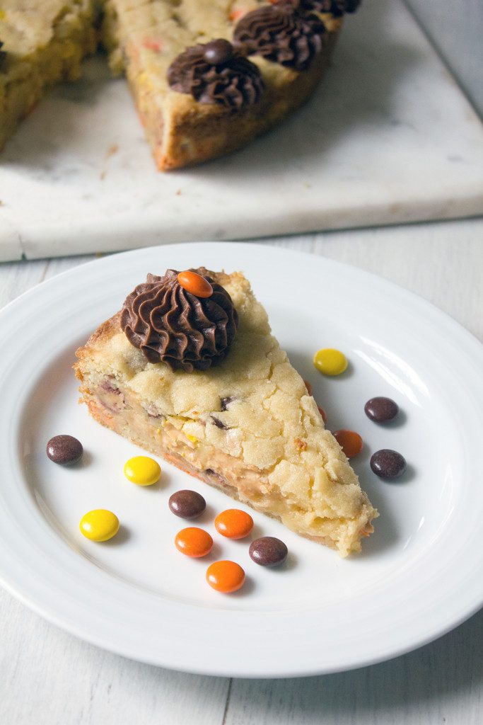 Head-on view of a slice of Reese's Pieces cookie cake on a white plate with Reese's Pieces with the rest of the cookie cake in the background