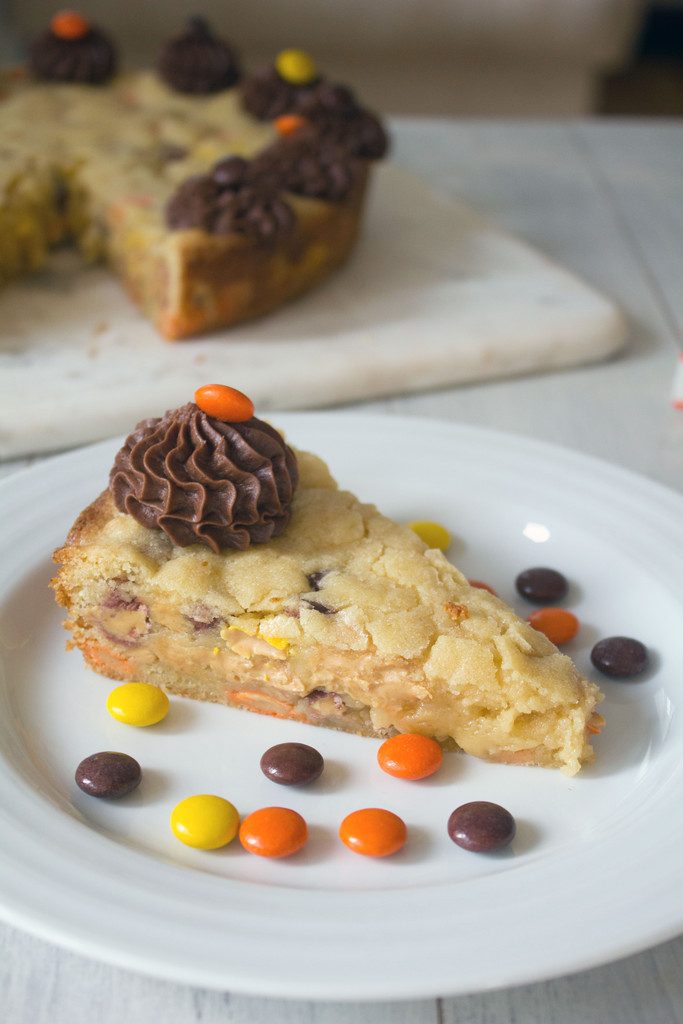 Head-on closeup view of a slice of Reese's Pieces cookie cake with Reese's Pieces on a white plate with remaining cookie cake in background