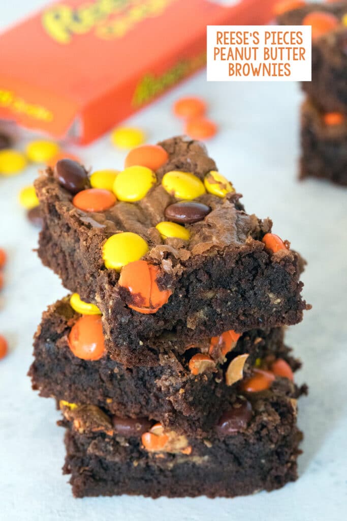 Head-on view of a stack of three Reese's Pieces peanut butter brownies with box of Reese's Pieces in the background with recipe title at top