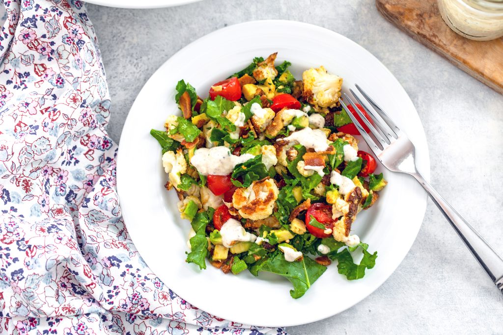 Overhead landscape view of a white bowl of roasted cauliflower BLT salad with a fork and flowered tea towel