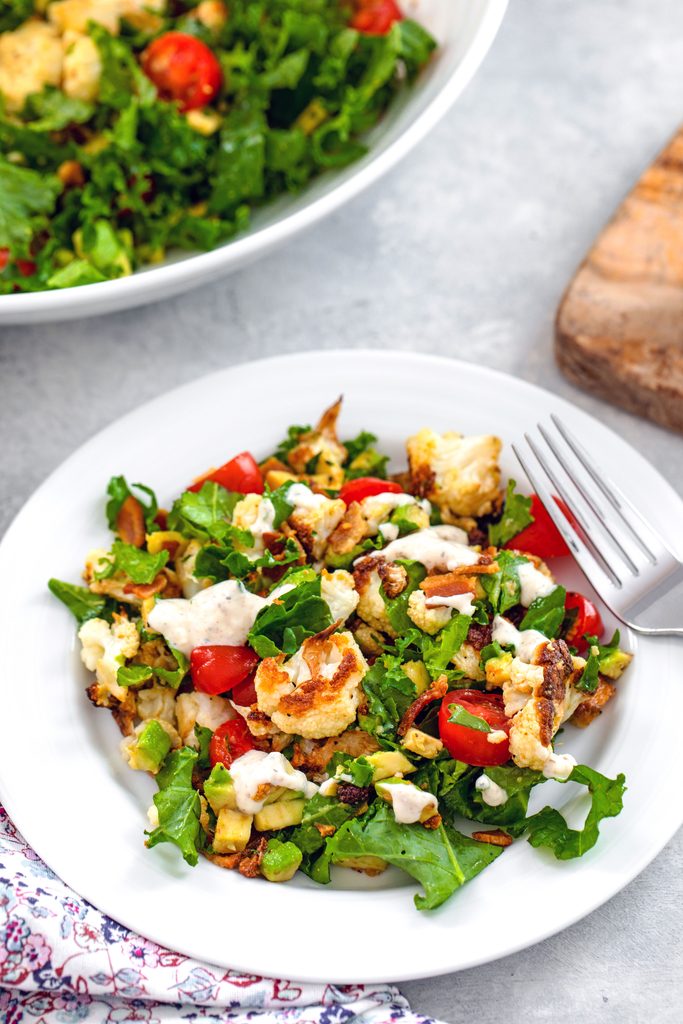 Overhead closeup view of a white bowl of roasted cauliflower BLT salad with a large bowl in the background