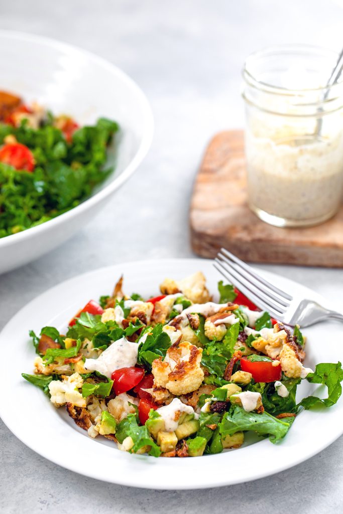 Head-on view of white bowl of roasted cauliflower BLT salad with a larger bowl in the background and cutting board with mason jar of creamy lemon dressing on it