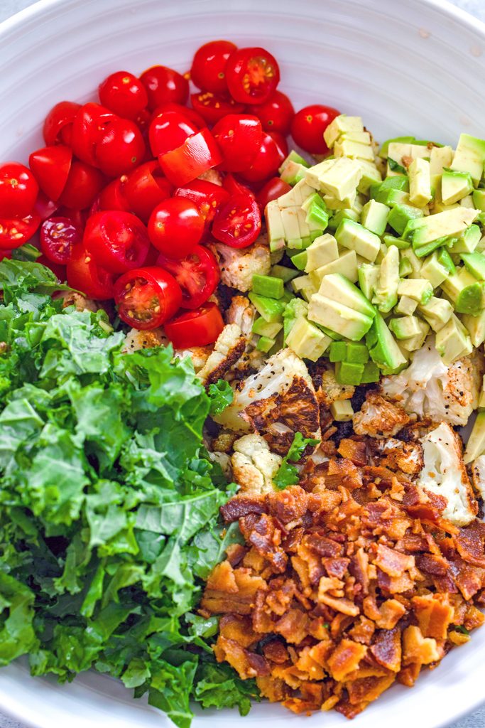 Overhead view of ingredients for roasted cauliflower BLT salad in a bowl including roasted cauliflower, chopped kale, cherry tomatoes, diced avocado, and crumbled bacon