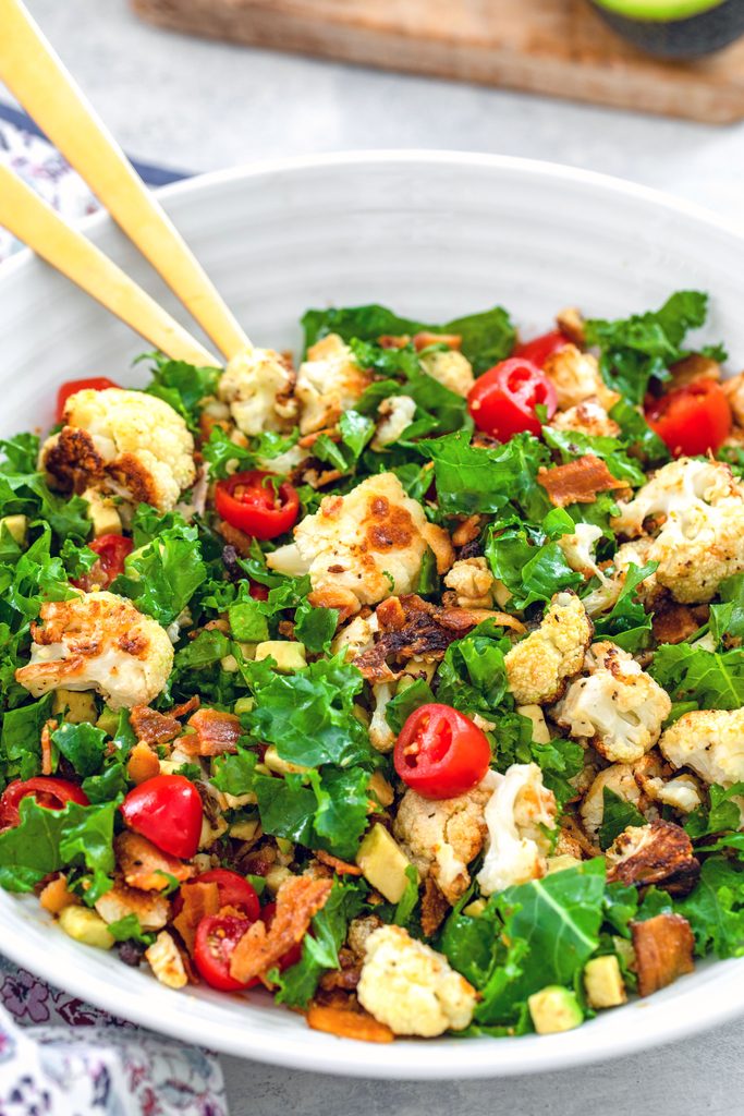 Head-on view of a large serving bowl of roasted cauliflower BLT salad with gold serving pieces