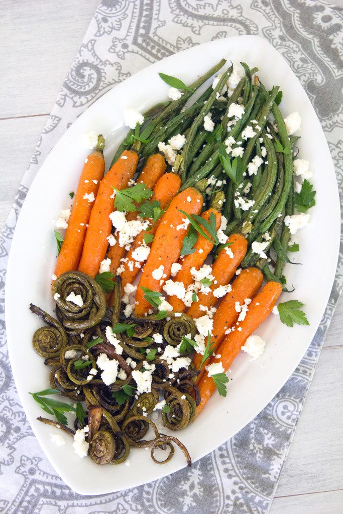 Bird's eye view of white platter with roasted fiddleheads, carrots, and haricot vert with feta cheese and parsley on a grey and white napkin