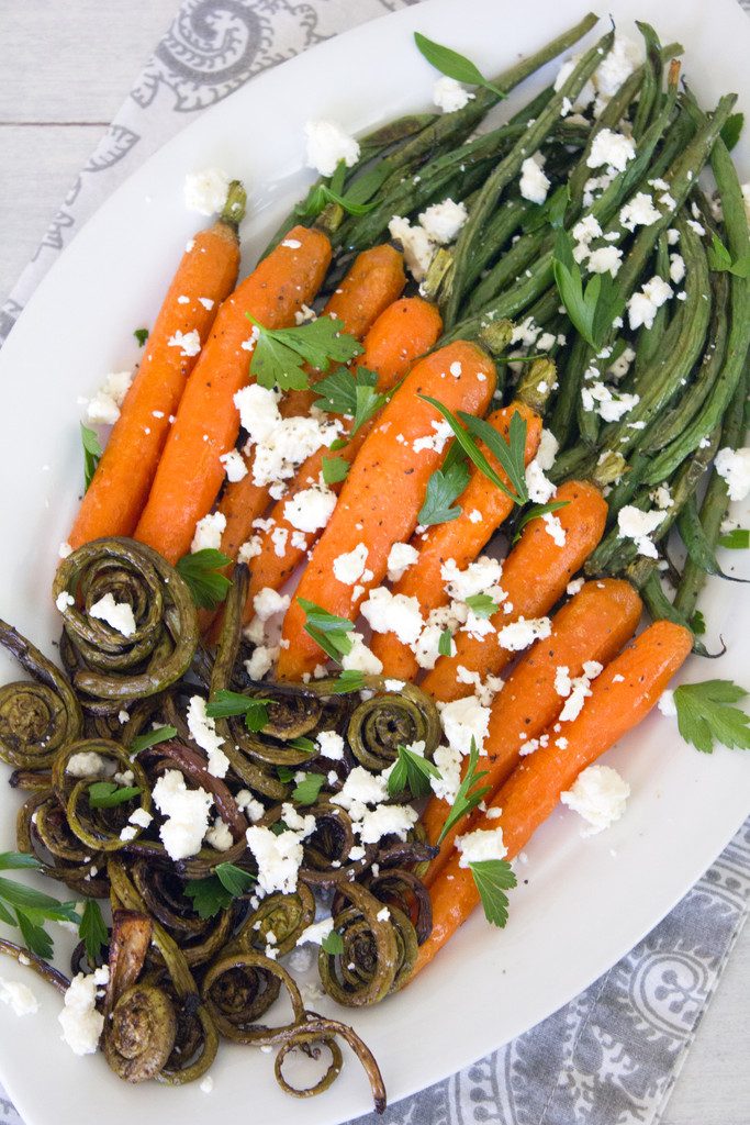 Overhead view of white platter with roasted fiddleheads, carrots, and haricot vert topped with feta cheese and parsley