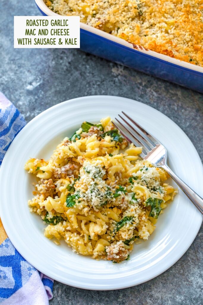 overhead view of a plate of roasted garlic mac and cheese with sausage and kale with a fork and serving dish in the background and recipe title at top