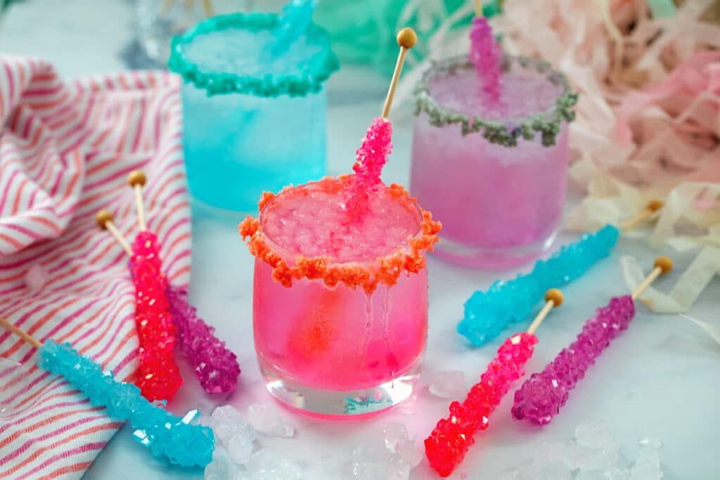 Landscape head-on view of a pink, blue, and purple rock candy cocktail with rock candy sticks all around