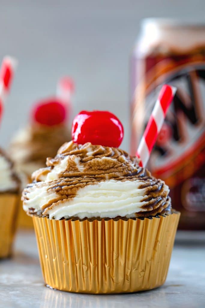 Close-up head-on photo of root beer float cupcake with red and white striped straw and cherry with can of root beer in the background