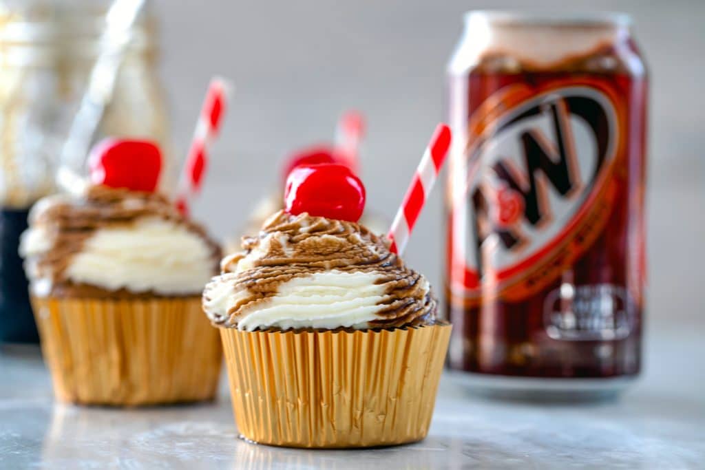 Landscape photo of root beer float cupcakes with red and white straws and cherries with jar of root beer syrup and can of root beer in the background