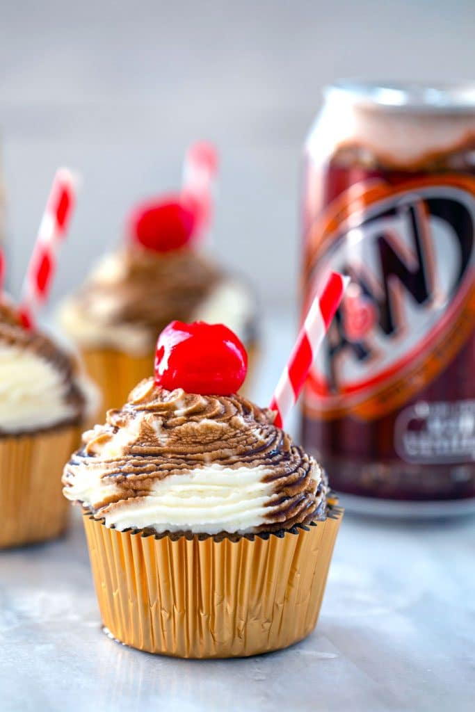 Head-on view of root beer float cupcake with red and white striped straw and cherry with other cupcakes and a can of root beer in the background