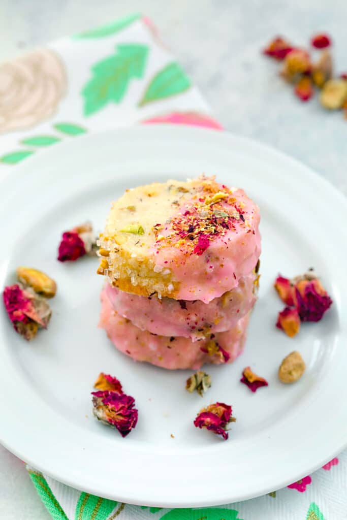 Head on view of rose pistachio cookies on a white plate with rose buds scattered around