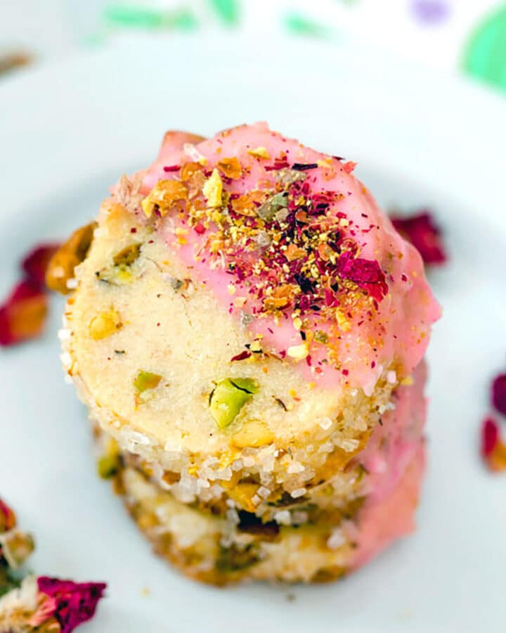 Closeup view of a stack of rose pistachio cookies with pink icing and crushed pistachios with rose petals all around