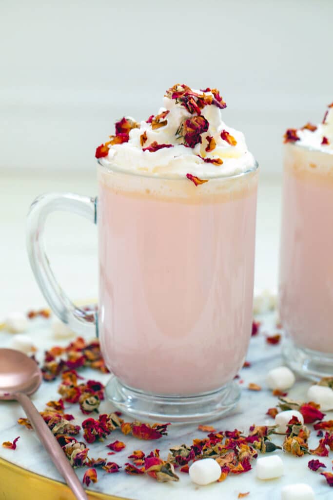 Head-on view of a big mug of pink colored rose white hot chocolate topped with whipped cream, mini marshmallows, and rose petals with second mug in background and rose petals all around