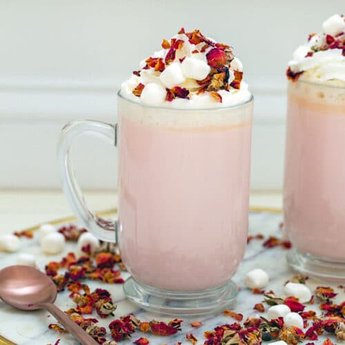 Head-on view of a large mug of pink colored rose white hot chocolate topped with whipped cream and rose petals with rose petals all around